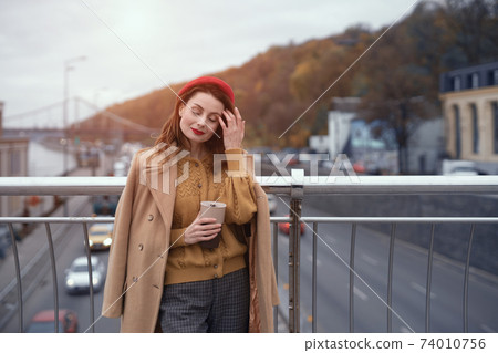 Woman with coffee mug on city street. Charming woman enjoying her cup of coffee standing in a spring urban bridge with traffic behind. French woman in red beret with background of urban city Woman with coffee mug on city street. Charming woman enjoying her cup of coffee standing in a spring urban bridge with traffic behind. French woman in red beret with background of urban city 74010756
