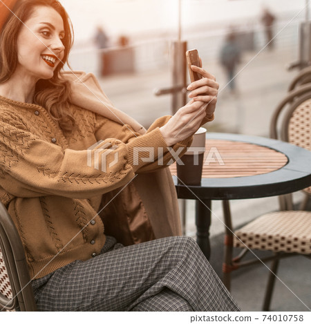 Beautiful young woman video calling using her smartphone while having cup of coffee sitting in a spring outdoor cafe. French woman in red beret with background of urban city. Square cropped Beautiful young woman video calling using her smartphone while having cup of coffee sitting in a spring outdoor cafe. French woman in red beret with background of urban city. Square cropped 74010758