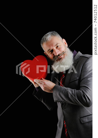 Mature bearded man wear suit holds red heart shaped gift box, isolated black background 74012512