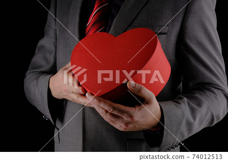 Unrecognizable man in suit holds out heart shaped gift red box, congratulatory concept, black background 74012513