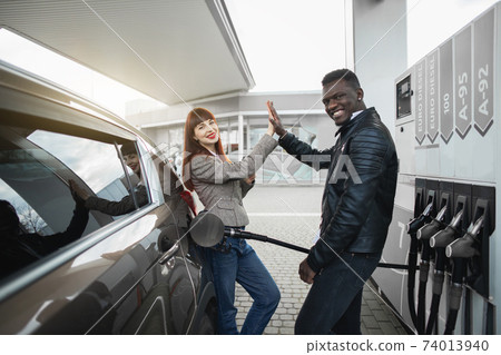 Couple at gas station. Happy joyful multiracial couple of friends or colleagues, posing to camera, having fun while refueling luxury car at petrol station, giving high five each other and smiling Couple at gas station. Happy joyful multiracial couple of friends or colleagues, posing to camera, having fun while refueling luxury car at petrol station, giving high five each other and smiling 74013940