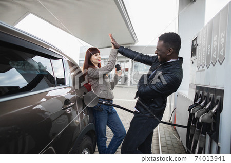 Couple at gas station. Happy joyful multiracial couple of friends or colleagues, having fun while refueling luxury car at petrol station, giving high five each other and smiling 74013941