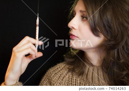Vaccine concept. Female portrait of caucasian girl holding a syringe and looking at it on black background in studio. 74014263