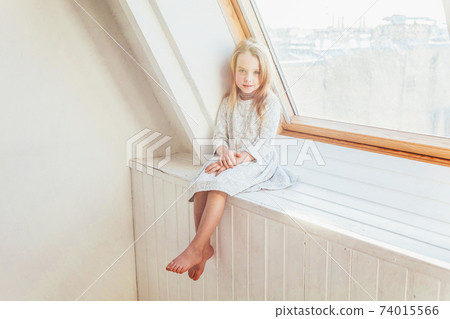 Little cute sweet smiling girl in white dress sitting on window sill in bright light living room at home indoors. Childhood schoolchildren youth relax concept. 74015566
