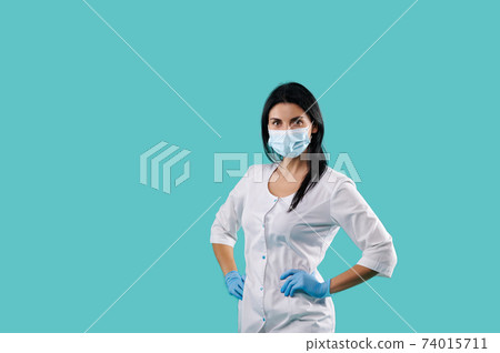 Portait on a blue background with a copy space of a young female doctor wearing medical uniform, protective mask and latex blue gloves. Healthcare concept Portait on a blue background with a copy space of a young female doctor wearing medical uniform, protective mask and latex blue gloves. Healthcare concept 74015711