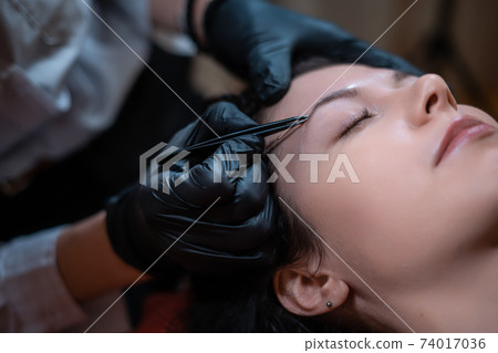 beautiful female at beautician on eyebrows correction procedure with tweezers beautiful female at beautician on eyebrows correction procedure with tweezers 74017036