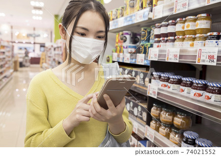A female shopper in a mask looking for products while looking at a smartphone at a supermarket 74021552