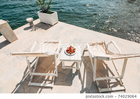 White wooden beach chairs by the sea. Wharf on the beach with beach furniture. A wooden table with a plate and strawberries. White wooden beach chairs by the sea. Wharf on the beach with beach furniture. A wooden table with a plate and strawberries. 74029064