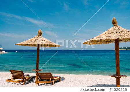 Two wooden loungers on the white sand, on a paradise beach near the town of Primosten in Croatia. Straw beach umbrella. Blue sky and azure Adriatic Sea. The concept of beach holidays and travel. Two wooden loungers on the white sand, on a paradise beach near the town of Primosten in Croatia. Straw beach umbrella. Blue sky and azure Adriatic Sea. The concept of beach holidays and travel. 74029216
