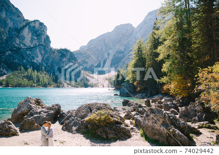 Bride and groom stand against the backdrop of stones overlooking Lago di Braies in Italy. Destination wedding in Europe, on Braies lake. Loving newlyweds walk against the backdrop of amazing nature. 74029442