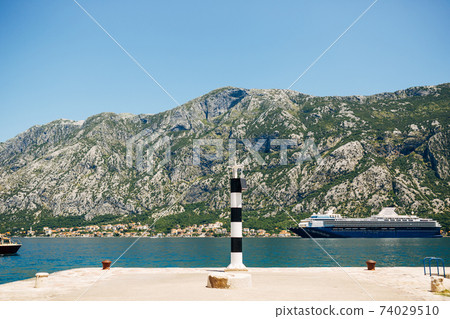A small striped metal lighthouse on the pier in Prcanj, Montenegro. Against the backdrop of rocky mountains and a huge cruise ship. A small striped metal lighthouse on the pier in Prcanj, Montenegro. Against the backdrop of rocky mountains and a huge cruise ship. 74029510