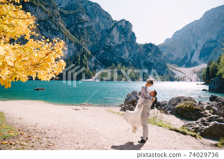 Bride and groom under an autumn tree, with fiery yellow foliage, at the Lago di Braies in Italy. Destination wedding in Europe, on Braies lake. The newlyweds in love are dancing and spinning. Bride and groom under an autumn tree, with fiery yellow foliage, at the Lago di Braies in Italy. Destination wedding in Europe, on Braies lake. The newlyweds in love are dancing and spinning. 74029736