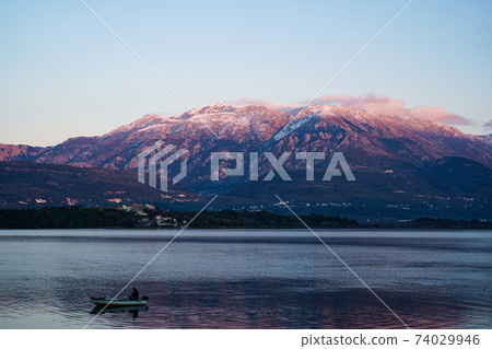 A fisherman is fishing in a boat in the Bay of Kotor, against the backdrop of the snow-covered Lovcen mountain at sunset. A fisherman is fishing in a boat in the Bay of Kotor, against the backdrop of the snow-covered Lovcen mountain at sunset. 74029946