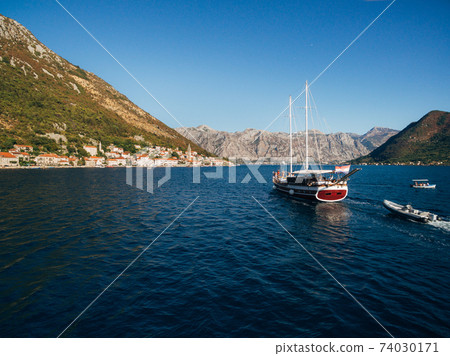 A large wooden sailing yacht sails against the backdrop of the city of Perast in Montenegro. A large wooden sailing yacht sails against the backdrop of the city of Perast in Montenegro. 74030171