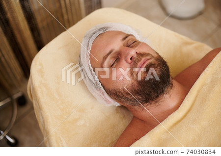 Young bearded handsome man lying on the massage table at beauty salon ready to receive spa treatment and beauty therapy 74030834