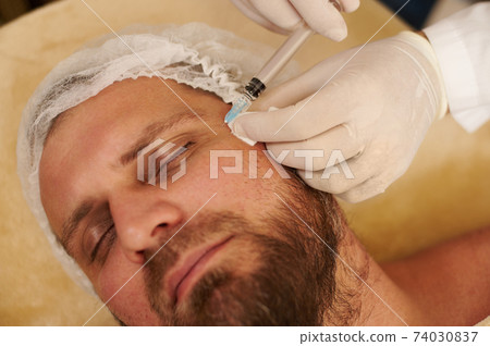 Focus on hands in medical gloves applying beauty injection for handsome bearded man in beauty salon. Rejuvenation concept 74030837