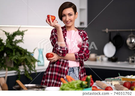 Holding bell pepper and tomato in the hand housewife wearing plaid shirt with a short hairstyle cooking vegetable ragout at new kitchen in new house. Healthy food at home Holding bell pepper and tomato in the hand housewife wearing plaid shirt with a short hairstyle cooking vegetable ragout at new kitchen in new house. Healthy food at home 74031702