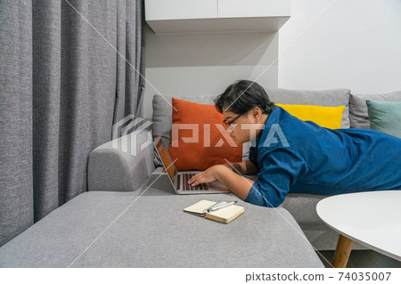 Woman lying prone on couch and working on laptop Woman lying prone on couch and working on laptop 74035007