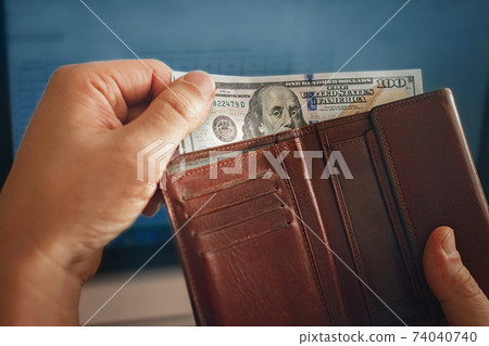 one hundred dollar banknotes in bundle protruded from leather brown wallet in men's hand on background blue laptop screen one hundred dollar banknotes in bundle protruded from leather brown wallet in men's hand on background blue laptop screen 74040740