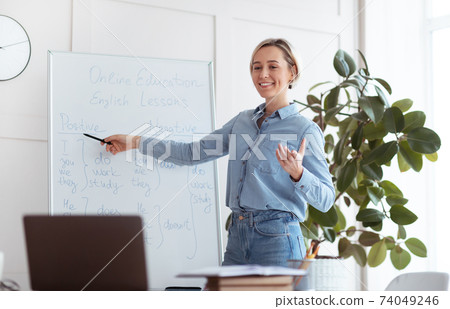 Joyful young teacher standing near blackboard, explaining grammar, giving online lesson to students on laptop 74049246
