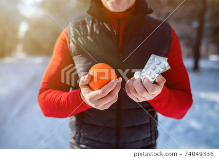 Healthy lifestyle vs medical treatment. Closeup of senior sportsman holding orange and pills at snowy winter park 74049336