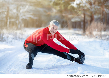 Outdoor winter activities concept. Joyful senior man stretching his legs before jogging at snowy park on cold day Outdoor winter activities concept. Joyful senior man stretching his legs before jogging at snowy park on cold day 74049486