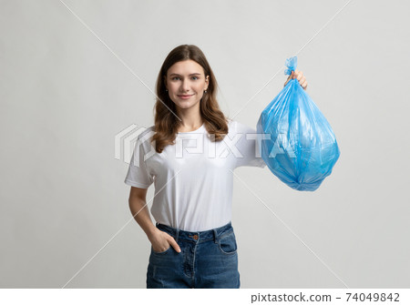 Happy young woman holding blue plastic garbage bag, standing on grey background 74049842
