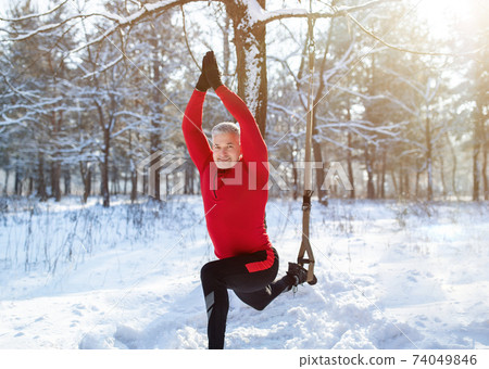 Outdoor winter fitness concept. Sporty senior man training with TRX resistance straps at snowy forest Outdoor winter fitness concept. Sporty senior man training with TRX resistance straps at snowy forest 74049846