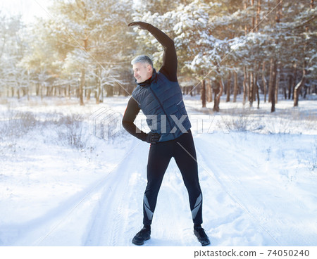 Outdoor seasonal sports. Full length portrait of positive mature guy doing exercises before jogging at snowy winter park 74050240