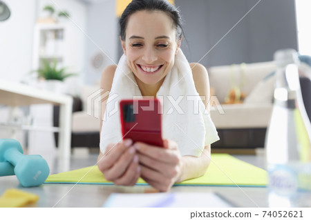 Woman in sportswear lying on gymnastic mat and looking on phone screen 74052621