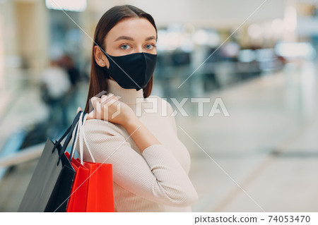 Positive young adult woman wearing protective medical face mask carrying paper shopping bags in hands at mall. Black friday and covid-19 new normal. Positive young adult woman wearing protective medical face mask carrying paper shopping bags in hands at mall. Black friday and covid-19 new normal. 74053470