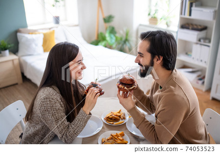 Young couple in love eating hamburgers indoors at home. Young couple in love eating hamburgers indoors at home. 74053523