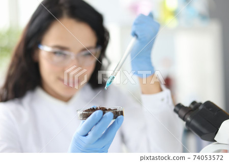 Female scientist dripping liquid from pipette into petri dish with soil 74053572