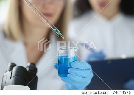 Female scientist dripping blue liquid from pipette into flask in laboratory 74053576