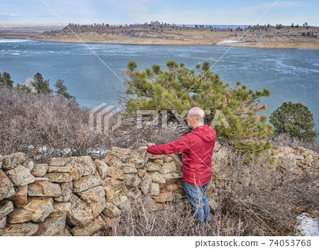 senior hiker at old stone fence overlooking frozen mountain lake 74053768