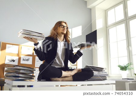 Calm worker holding papers and laptop and meditating in yoga pose on office desk Calm worker holding papers and laptop and meditating in yoga pose on office desk 74054310