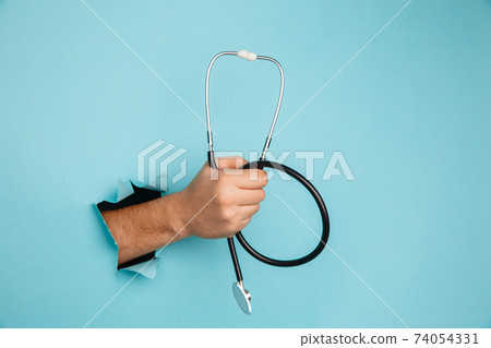 Stethoscope in a male hand on a blue background with a hole, medical concept 74054331