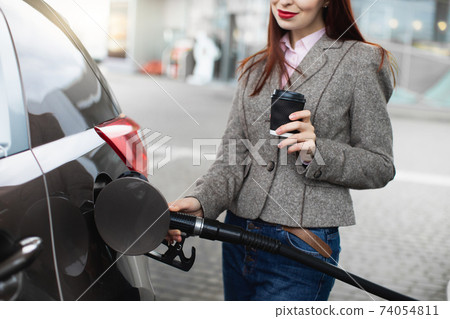 Cropped close up view of young woman holding take away coffee, having a break, standing near the car and refueling it on the gas station Cropped close up view of young woman holding take away coffee, having a break, standing near the car and refueling it on the gas station 74054811
