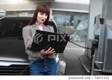 Close up portrait of beautiful Caucasian businesswoman, leaning on her car while refueling it at petrol station, working on laptop pc and smiling to camera Close up portrait of beautiful Caucasian businesswoman, leaning on her car while refueling it at petrol station, working on laptop pc and smiling to camera 74055105