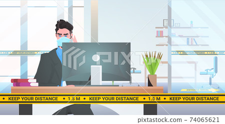 businessman in mask sitting at workplace desk with yellow tape for social distancing 74065621