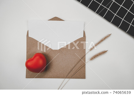 Blank white paper is placed on open brown paper envelope with red heart, bristly foxtail dry flower, black cloth on white background. Valentine's day concept Blank white paper is placed on open brown paper envelope with red heart, bristly foxtail dry flower, black cloth on white background. Valentine's day concept 74065639