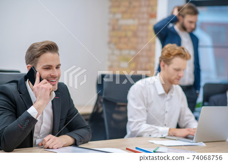 Smiling man talking on smartphone and working colleagues Smiling man talking on smartphone and working colleagues 74068756
