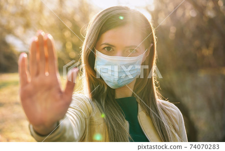 Young woman wearing blue disposable single use virus mouth nose mask, hold hand front of her as stop gesture, blurred sunset lit trees in background. Coronavirus covid-19 outbreak prevention concept Young woman wearing blue disposable single use virus mouth nose mask, hold hand front of her as stop gesture, blurred sunset lit trees in background. Coronavirus covid-19 outbreak prevention concept 74070283