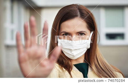 Young woman wearing white cotton virus mouth nose mask, holding hand in front of her as stop gesture, blurred building in background. Coronavirus covid-19 outbreak prevention concept Young woman wearing white cotton virus mouth nose mask, holding hand in front of her as stop gesture, blurred building in background. Coronavirus covid-19 outbreak prevention concept 74070284