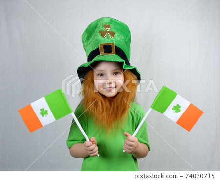 Saint Patricks Dayc elebration. Pretty smiling small girl with decorative red beard in green T-shirt and leprechaun's hat, with flags of Ireland in her hands Saint Patricks Dayc elebration. Pretty smiling small girl with decorative red beard in green T-shirt and leprechaun's hat, with flags of Ireland in her hands 74070715