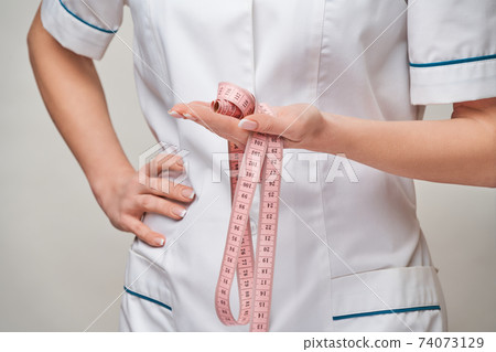 Cheerful woman doctor or nurse holding a measure tape standing over grey background 74073129