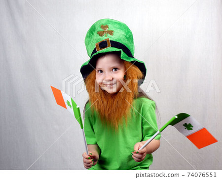 Saint Patricks Day holiday. Portrait of adorable smiling girl with decorative red beard, green clover leaf on her cheek and leprechaun hat, holds flags of Ireland in her hands 74076551