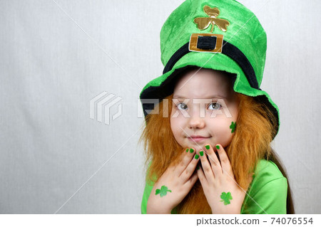 Saint Patricks Day celebration. Nice pensive little girl with decorative red beard, green shamrok leaf on her cheek and leprechaun hat, raised her hands with green nails and clover leaves to her face Saint Patricks Day celebration. Nice pensive little girl with decorative red beard, green shamrok leaf on her cheek and leprechaun hat, raised her hands with green nails and clover leaves to her face 74076554