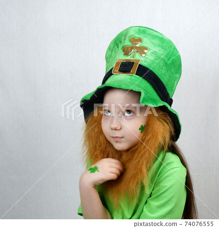 Saint Patricks Day holiday. Portrait of adorable pensive girl with clover leaf on her cheek and hand, decorative red beard in green clothes and leprechaun hat Saint Patricks Day holiday. Portrait of adorable pensive girl with clover leaf on her cheek and hand, decorative red beard in green clothes and leprechaun hat 74076555
