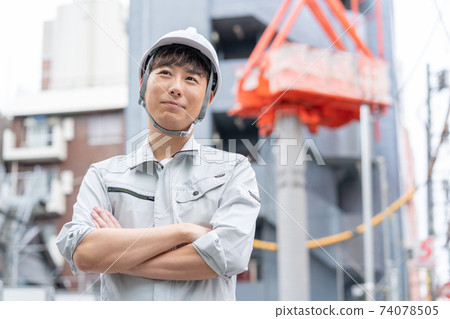 一名戴著頭盔和雙臂交叉在建築工地的年輕工人 74078505